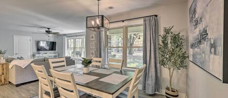 A dining area with a table, chairs, and a hanging light fixture. There is a television and a plant in the background.