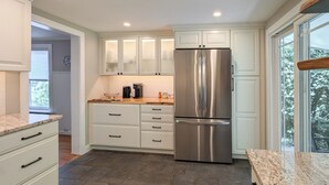 A modern kitchen with white cabinets, a stainless steel refrigerator, granite countertops, and a view through the window. - Charming Abode w/ Patio - Walk to Town + River! (Stockton)