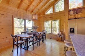 A wooden cabin interior with a dining table, chairs, a kitchen area, and large windows. - 12 Mi to Sugar Mountain: Luxe Banner Elk Escape (Banner Elk)