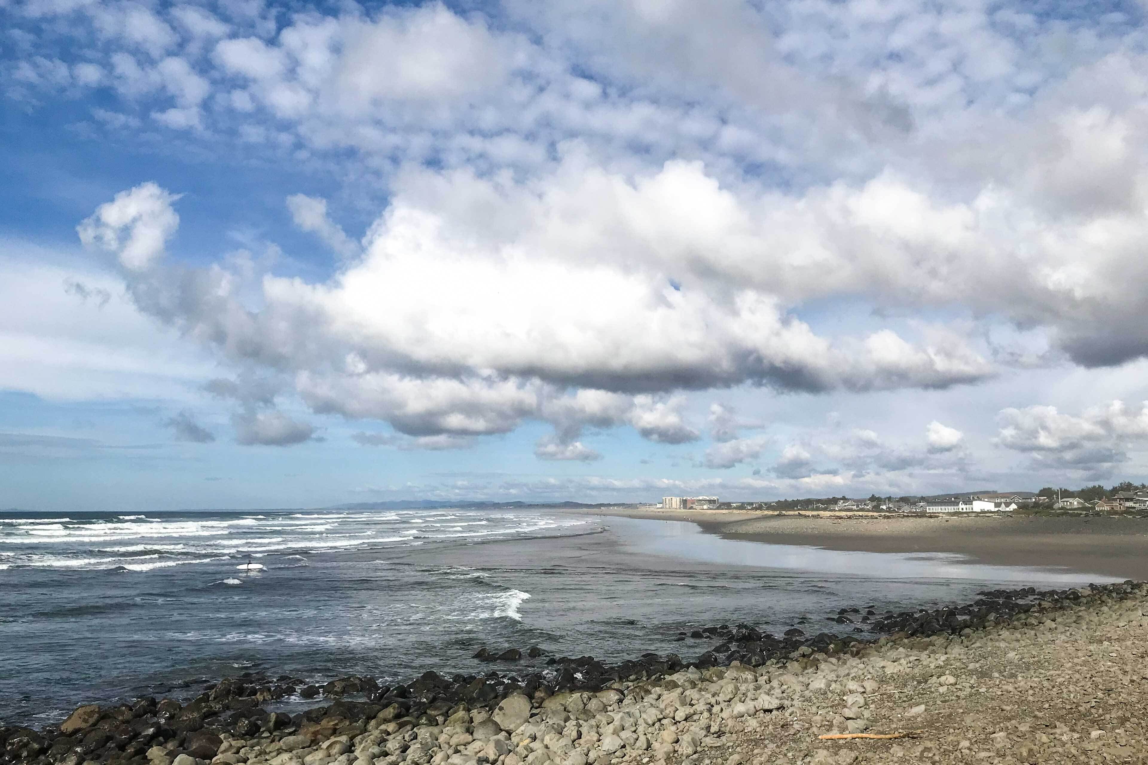A coastal beach scene with waves, a rocky shoreline, and buildings in the distance.