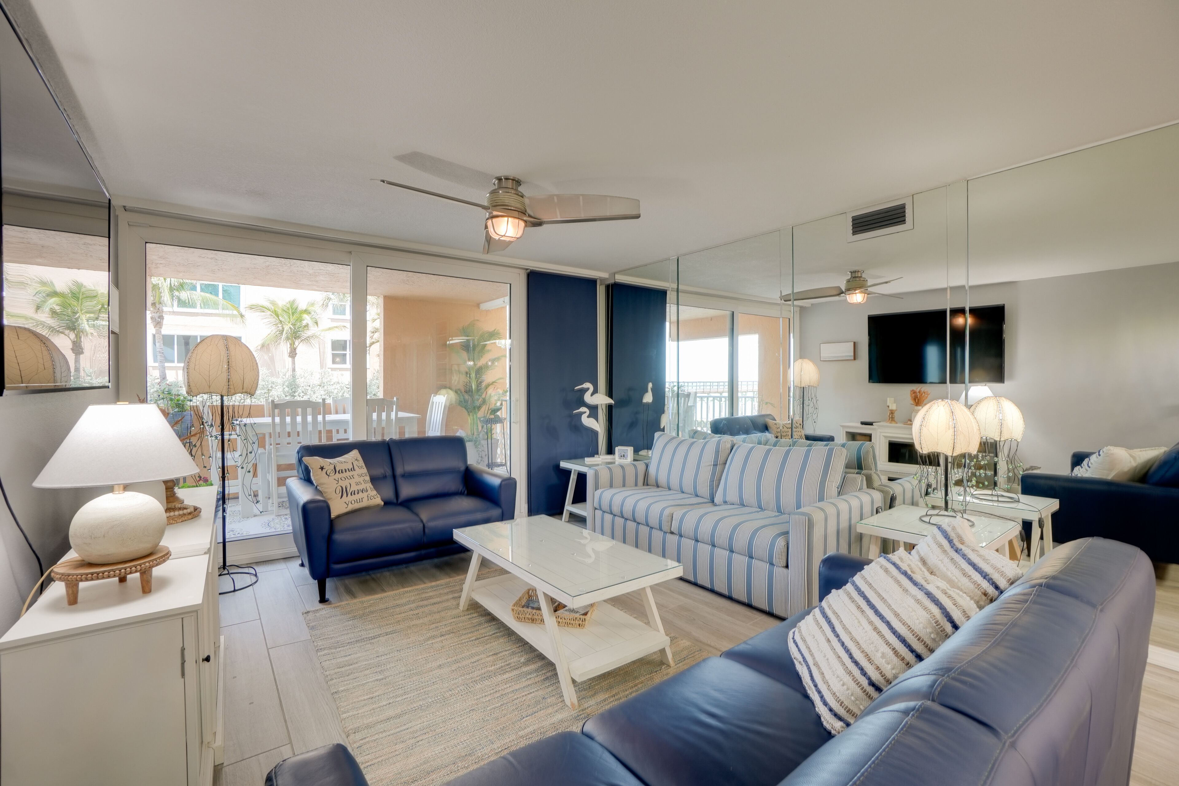 A modern living room with a blue sofa, striped armchair, and a glass coffee table. There is a ceiling fan, a TV mounted on the wall, and a large window with a view of palm trees.