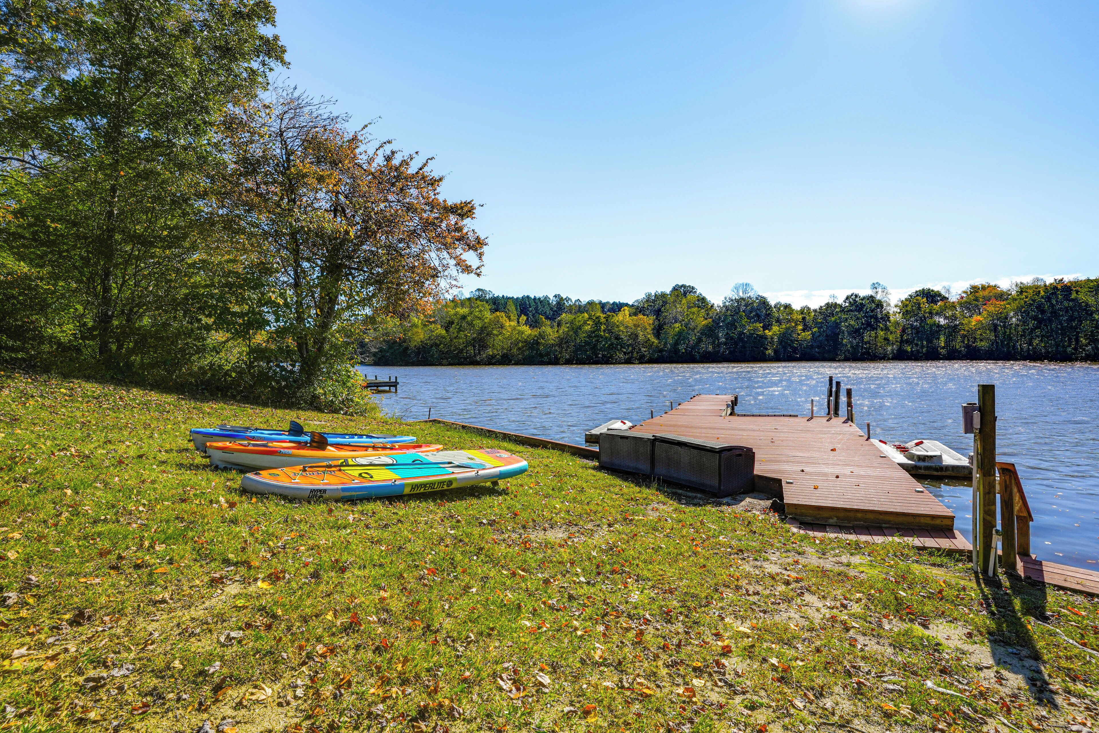 A grassy lakeside area with a wooden dock extending into the water, featuring colorful kayaks and a small boat.
