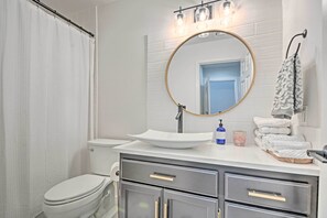 Une salle de bain avec un lavabo blanc, un miroir rond et des toilettes. On y trouve des serviettes et un distributeur de savon sur le comptoir.