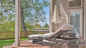 Eine überdachte Terrasse im Freien mit zwei Liegestühlen und einem Tisch, einem weißen Sonnenschirm und Blick auf ein Haus und Bäume.