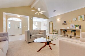 A spacious living room with a glass coffee table, a beige sofa, and a white armchair. There is a ceiling fan, a chandelier, and a wall-mounted clock. - Chic Home w/ Club Access Near Bay St Louis! (Diamondhead)