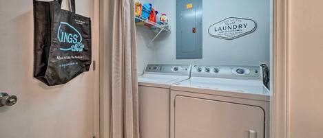 A laundry room with a washing machine, a dryer, and a shelf with cleaning supplies. A black tote bag hangs on the door.