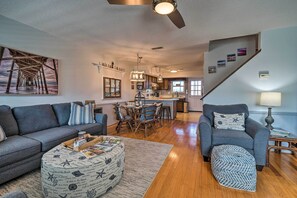A living room with a sofa, ottoman, and a dining area with chairs and a table. - Holden Beach Vacation Rental: Short Path to Beach! (Supply)