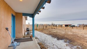 A covered porch with a blue door, a shovel leaning against the wall, and a view of a rural landscape with buildings in the distance. - Ideally Located Taos Vacation Rental w/ Gas Grill (Ranchos De Taos)
