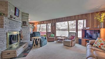A cozy living room with a fireplace, comfortable seating, a television, and large windows overlooking a body of water.
