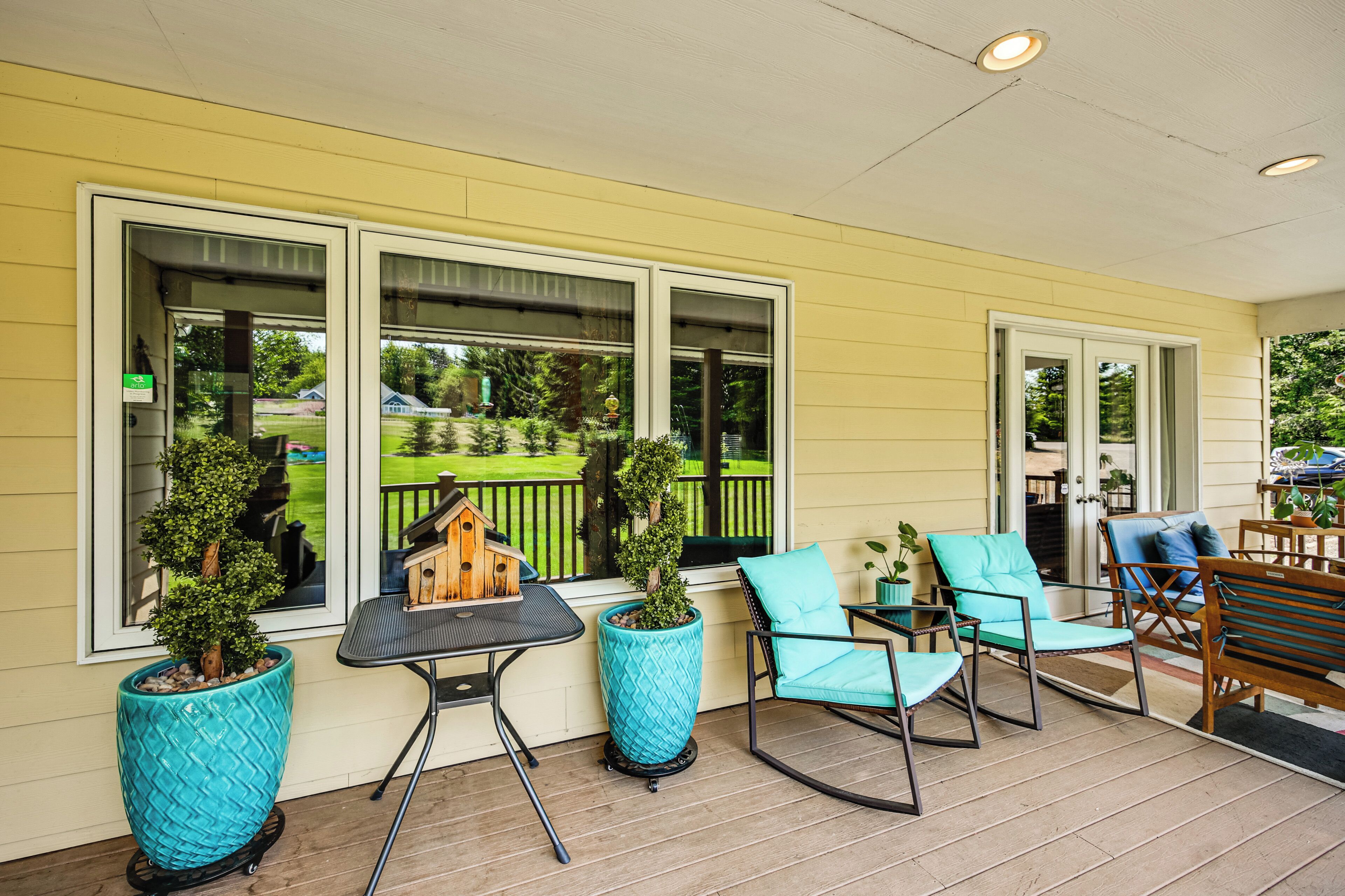 A covered porch with a yellow exterior, large windows, and outdoor furniture including chairs and a table.