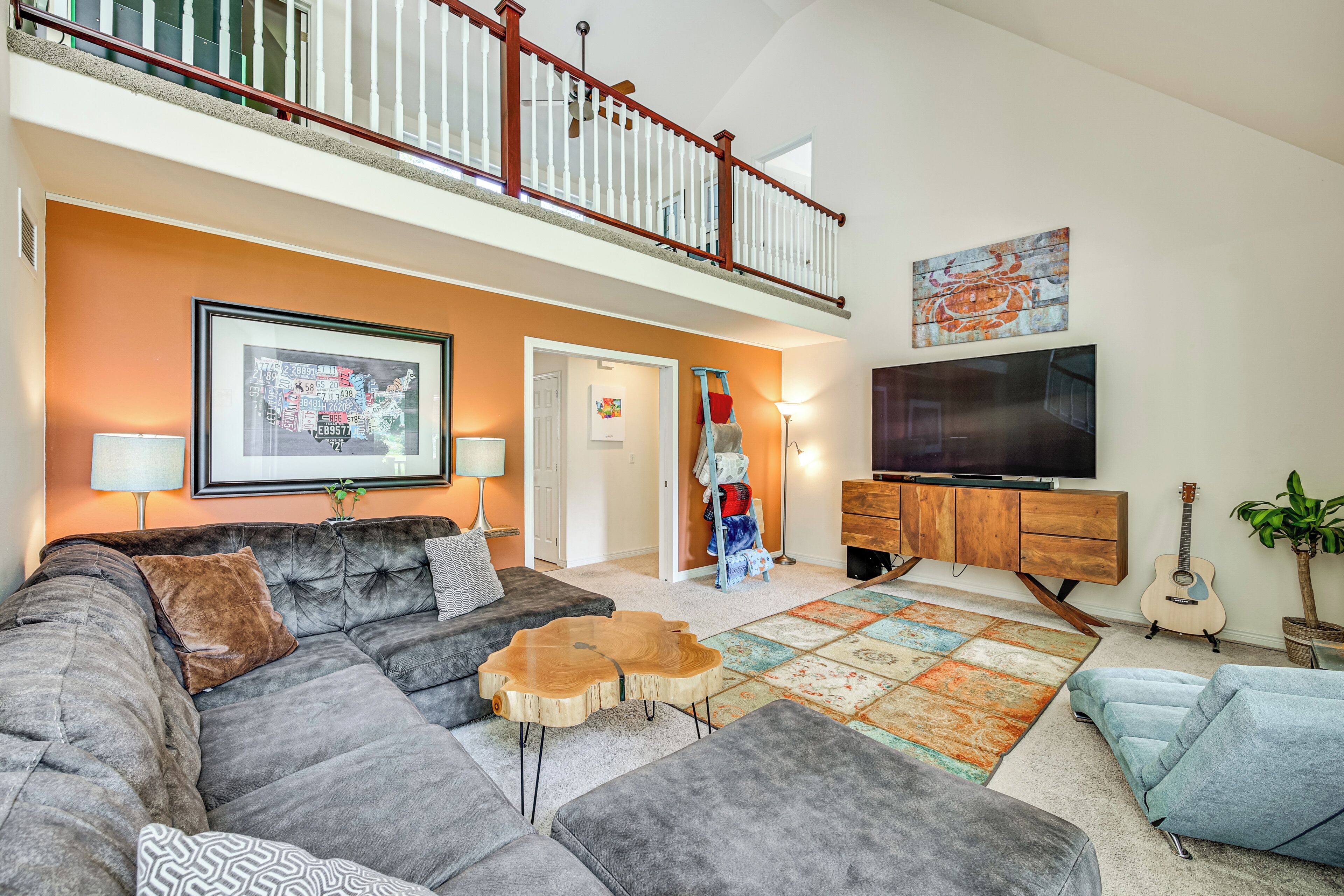 A spacious living room with a large sectional sofa, a wooden coffee table, and a colorful rug. A television is mounted on the wall, and there is a staircase leading to an upper level.