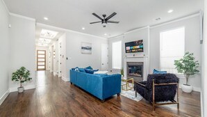 A spacious living room with hardwood flooring, a blue sofa, a black furry chair, a fireplace, and a ceiling fan. - Glam Paradaise 5BRs Home in Downtown Dallas (Dallas)