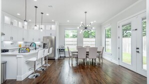 A modern kitchen with white cabinets, a central island with bar stools, a dining area with a table and chairs, and large windows offering a view of the outdoors. - Glam Paradaise 5BRs Home in Downtown Dallas (Dallas)