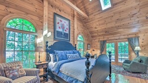 A spacious bedroom with a large bed, a sitting area with a chair, and multiple windows offering views of the outdoors.