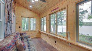 A cabin with a wooden interior, large windows, a ceiling fan, and a bench with patterned cushions. - Lakefront Maine Retreat w/ Deck, Dock & Fire Pit! (Greenville)