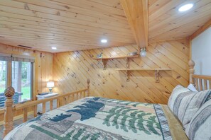 A wooden-paneled bedroom with a bed, a nightstand, and a window with a view of the outdoors. - Lakefront Maine Retreat w/ Deck, Dock & Fire Pit! (Greenville)