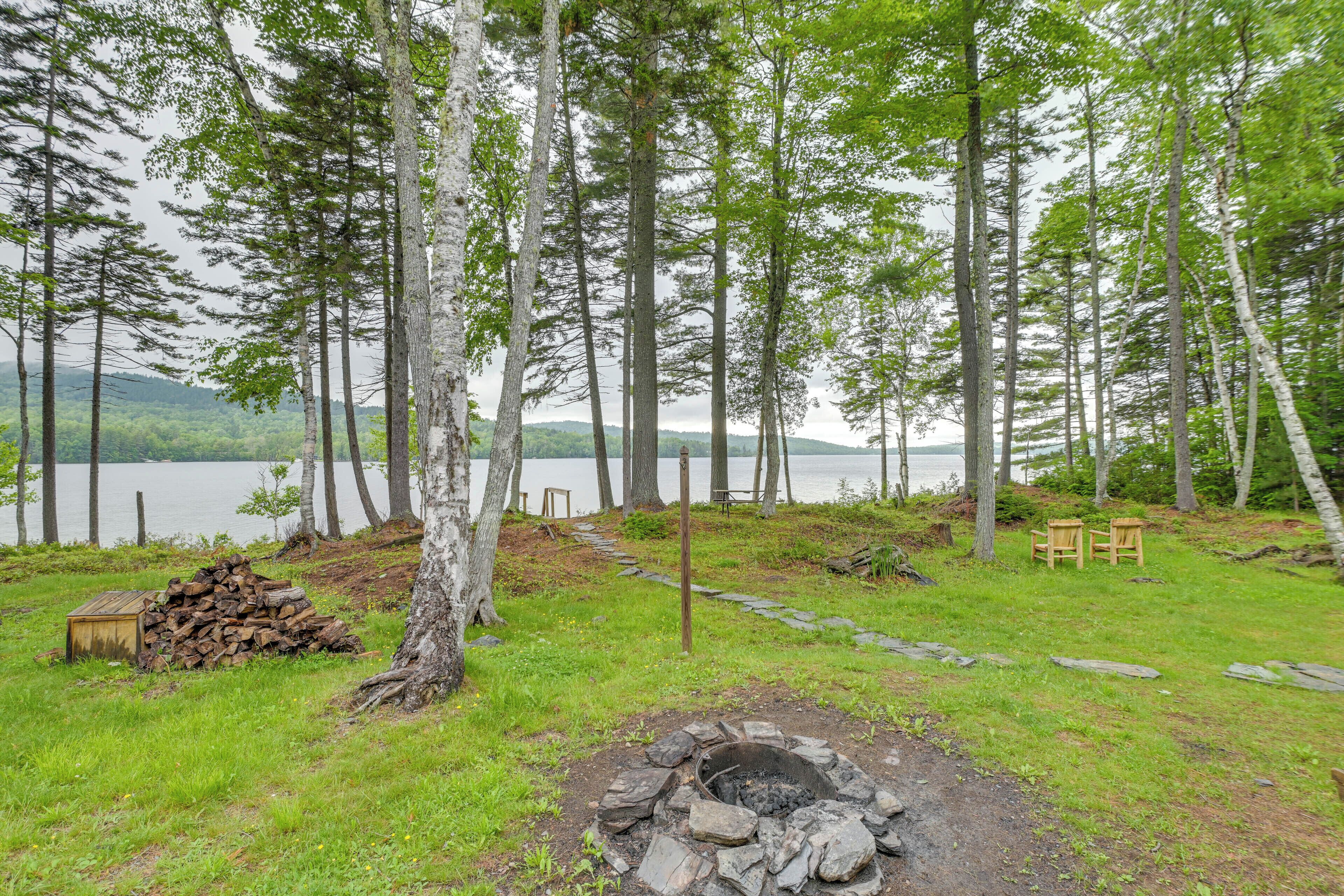 Un camping con un pozo de fuego, una pila de leña y un banco junto a un lago rodeado de árboles.