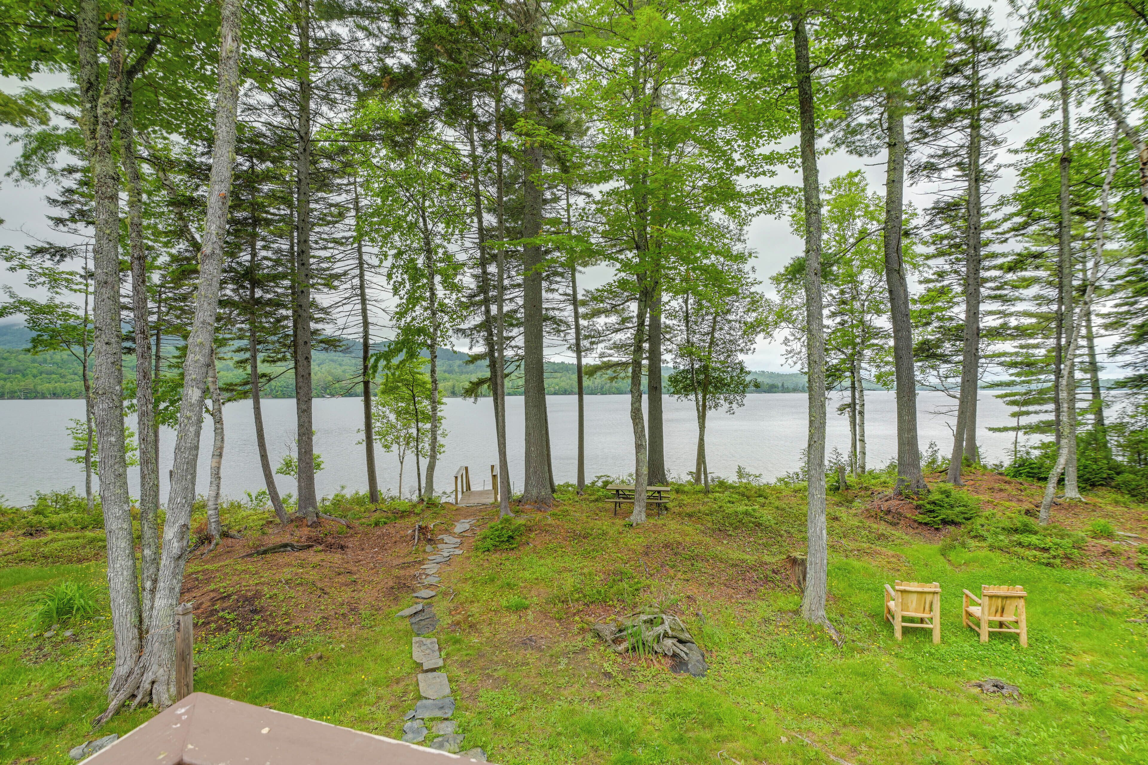 A lakeside view with tall trees, a stone path, and two wooden chairs.