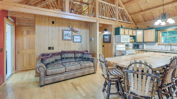 A spacious wooden interior with a high ceiling, a central wooden dining table surrounded by rustic chairs, a sofa with patterned cushions, and a kitchen area with green cabinets and a window overlooking greenery. - Lakefront Maine Retreat w/ Deck, Dock & Fire Pit! (Greenville)