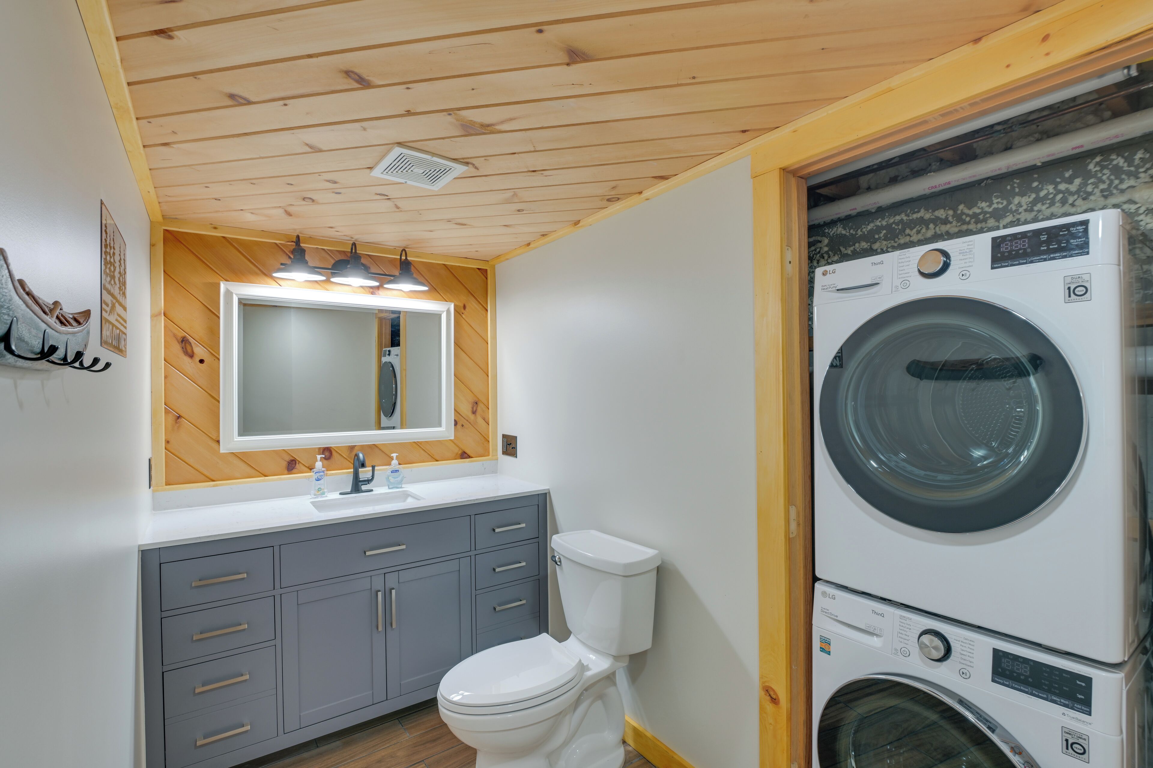 Un bagno compatto con soffitto in legno, un lavandino con specchio, un WC e una lavatrice e asciugatrice affiancate.