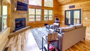 A cabin interior with a stone fireplace, wooden floors, a sofa, and large windows. - Family-friendly Murphy Cabin w/ Fire Pit! (Murphy)
