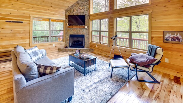 A cozy cabin interior with a stone fireplace, wooden walls, and large windows offering views of the forest. - Family-friendly Murphy Cabin w/ Fire Pit! (Murphy)