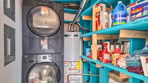 A laundry area with two washing machines, a water heater, and shelves stocked with cleaning supplies and towels.