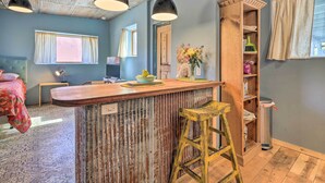 A room with a wooden bar counter, two hanging pendant lights, a wooden stool, a trash can, and a window with curtains.
