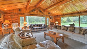 A log cabin interior with a cozy living area featuring a sofa, armchairs, and a coffee table. Large windows provide a view of the surrounding forest.