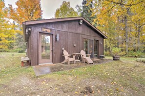 A rustic cabin with a wooden exterior, a door, and a small porch area with two Adirondack chairs and a table.