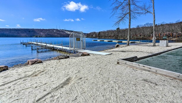 Une plage de sable avec une jetée qui s’avance dans un lac, entourée d’arbres et d’une colline lointaine.