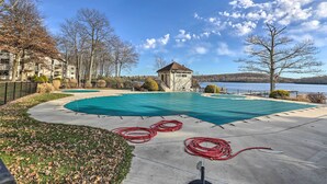 Une piscine recouverte d’une bâche bleue, entourée d’une terrasse en béton, d’arbres, et offrant une vue sur un lac et des bâtiments en arrière-plan.