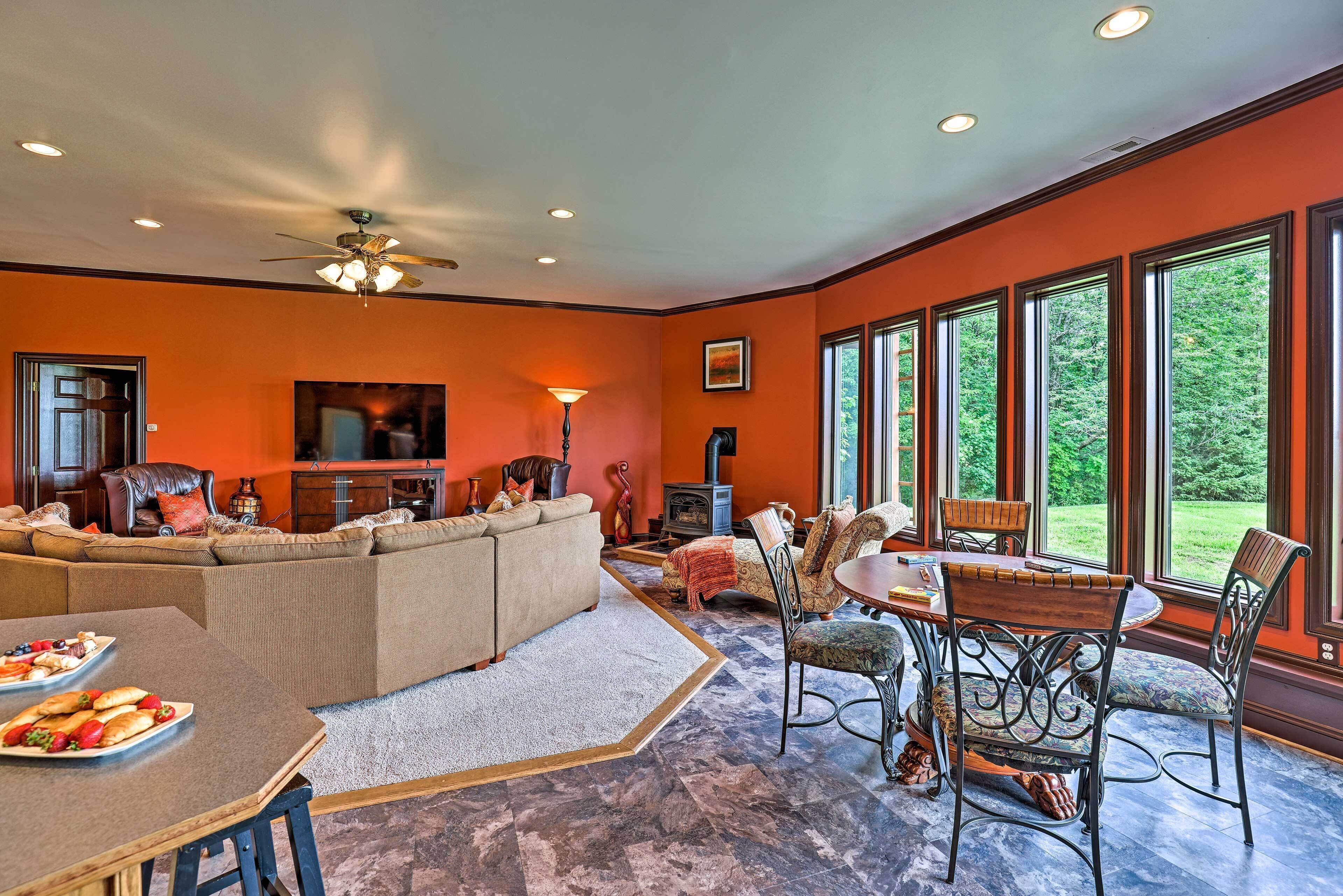 A spacious living room with a large sofa, dining table, and chairs. There is a fireplace, a television, and large windows with a view of greenery outside.