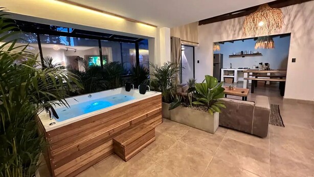 A modern living room with a wooden bathtub, large windows, and a dining area with a kitchen in the background. - Increible Casa con Jacuzzi en Poblado-lleras (Medellín)