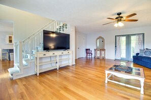 A spacious living room with hardwood flooring, a white entertainment unit, a flat-screen TV, a ceiling fan, a blue couch, and a glass coffee table. - Spacious White Rivers Jewel: Fishing Haven! (Flippin)