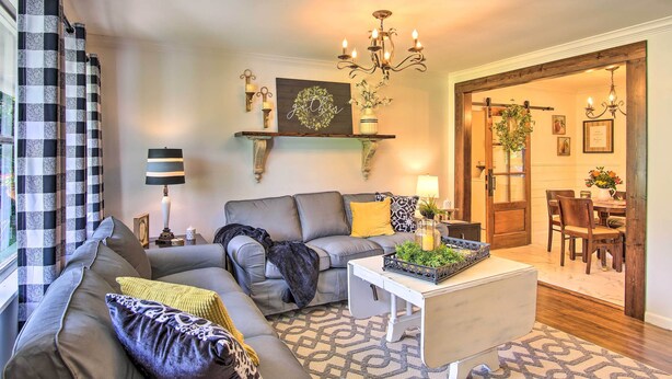 A living room with a sofa, coffee table, and decorative items. There is a chandelier, a wall-mounted shelf, and a door leading to another room. - Charming Country Cottage: 5 Mi to Downtown Tulsa! (Tulsa)
