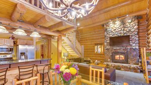 L’intérieur d’une cabane en rondins est agrémenté d’une cheminée en pierre, d’un plafond en bois et d’un coin repas avec une table et des chaises en verre.