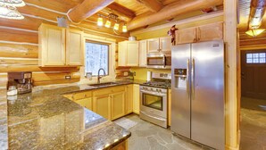 Une cuisine rustique aménagée dans une cabane en rondins, avec des meubles en bois, des plans de travail en granit, des appareils électroménagers en acier inoxydable et une grande fenêtre.