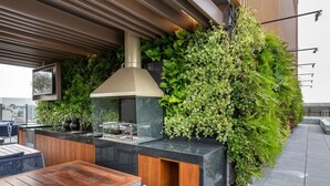 Cucina esterna con barbecue integrato, tavolo e sedie in legno, circondata da piante verdi rigogliose.
