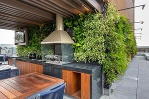 An outdoor kitchen with a built-in grill, wooden table, and chairs, surrounded by lush green plants.