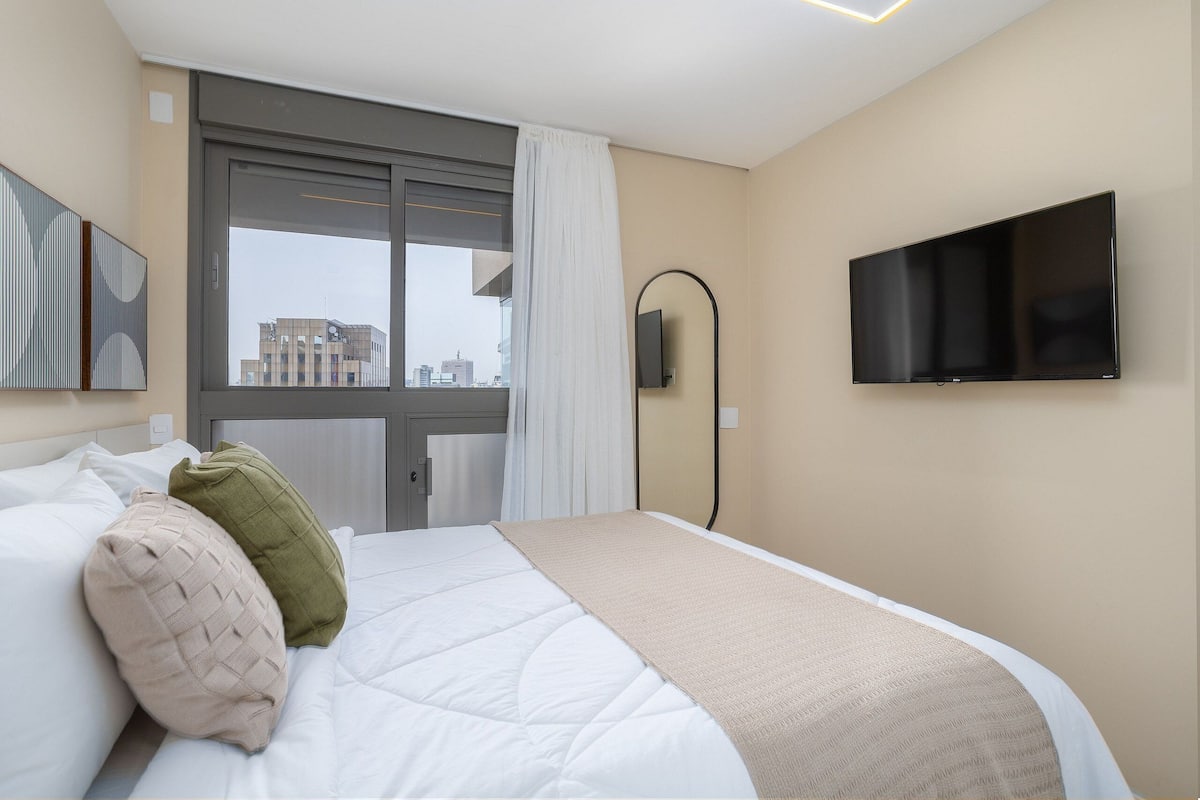 Une chambre d’hôtel avec un lit, une télévision, une fenêtre avec des rideaux et une vue sur des immeubles.