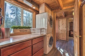 L’intérieur d’un chalet en bois, avec un lave-linge, une pancarte « Bienvenue » et une vue sur la forêt à travers de grandes fenêtres.