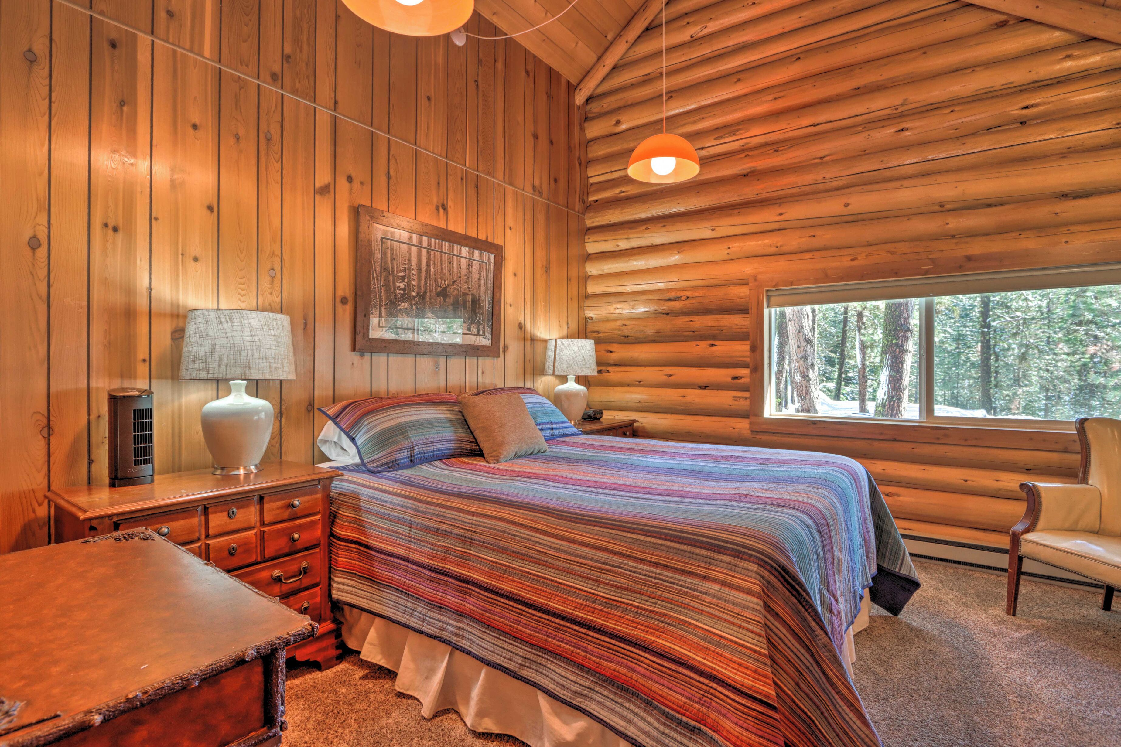 Quarto de cabana de tronco com cama, duas mesinhas de cabeceira, uma cadeira e uma janela com vista para a floresta.