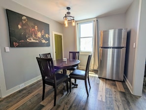 Un coin repas avec une table ronde et quatre chaises, un réfrigérateur moderne en acier inoxydable, un tableau accroché au mur et une fenêtre avec des rideaux.