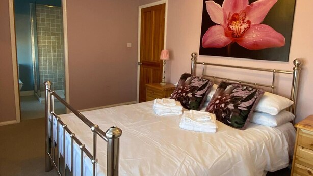 Een slaapkamer met een bed, nachtkastje, houten dressoir, badkamer met douche en een groot bloemschilderij aan de muur.