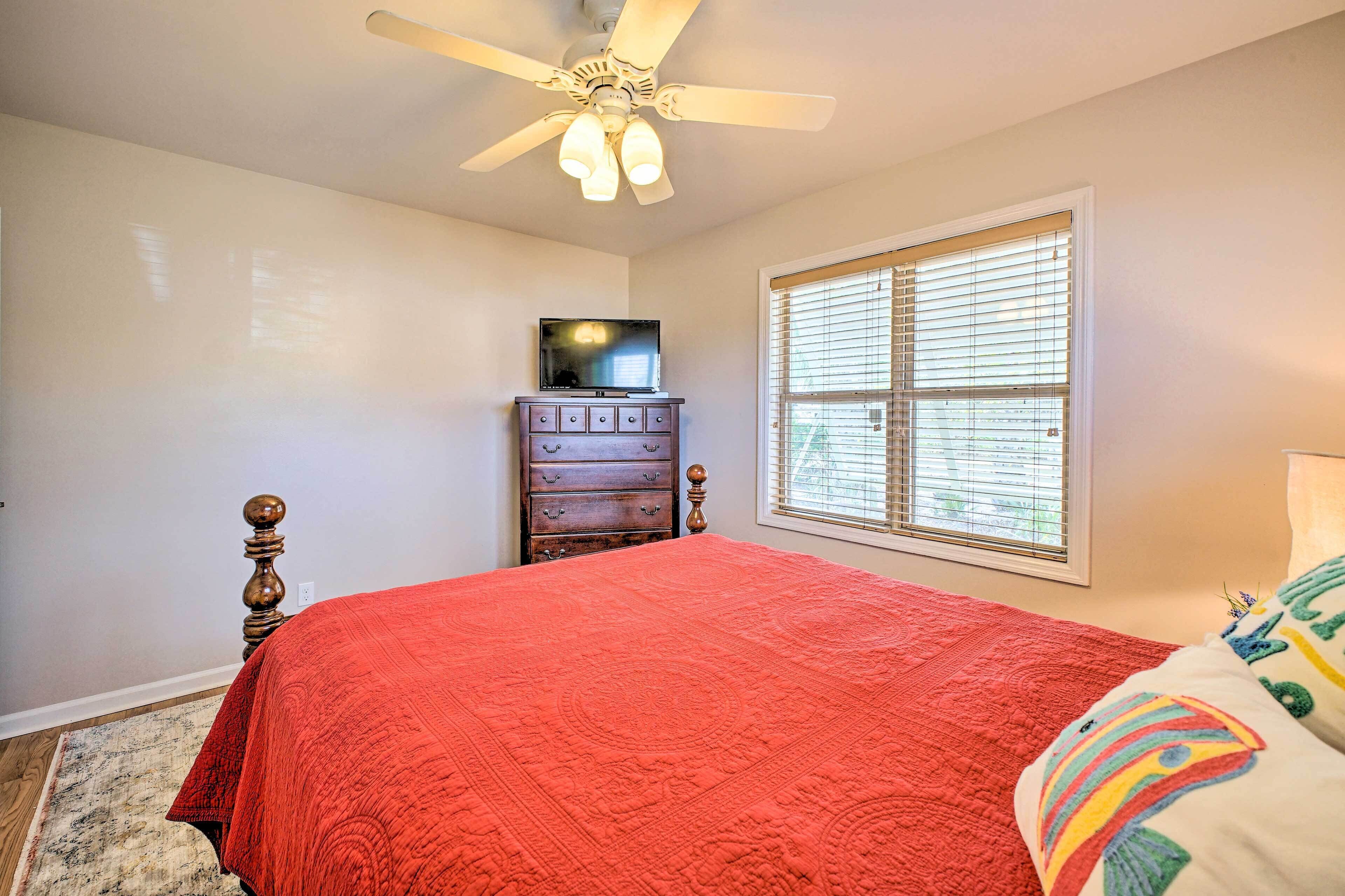 A bedroom with a red bedspread, a wooden dresser, a television on top, and a ceiling fan.