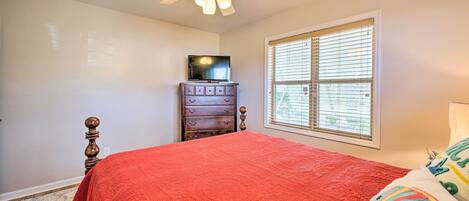 A bedroom with a red bedspread, a wooden dresser, a television on top, and a ceiling fan.