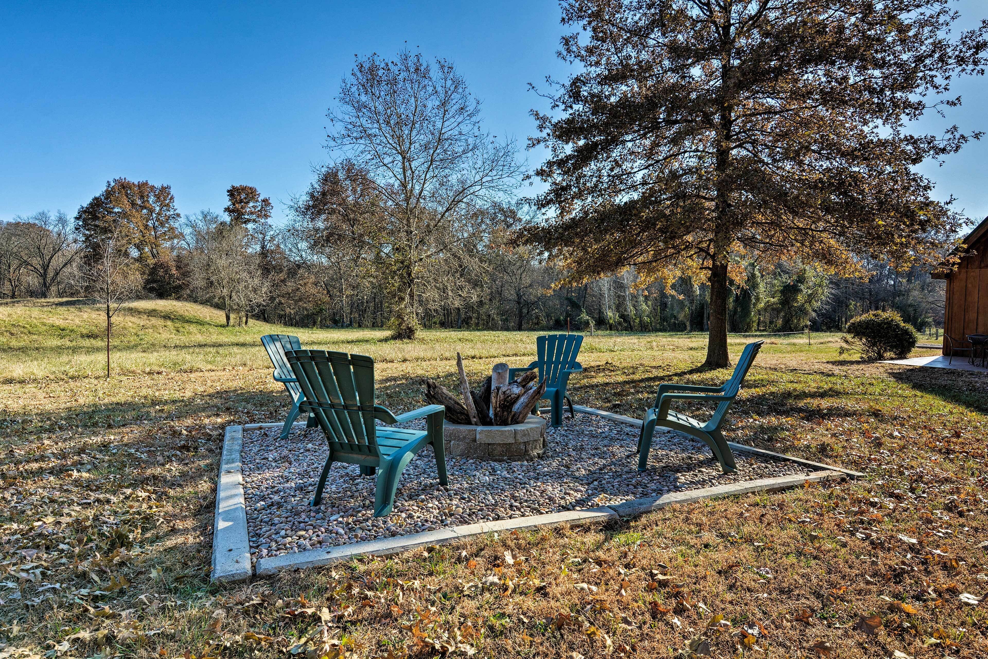 A fire pit with four green Adirondack chairs arranged around it, set on a gravel area with fallen leaves.
