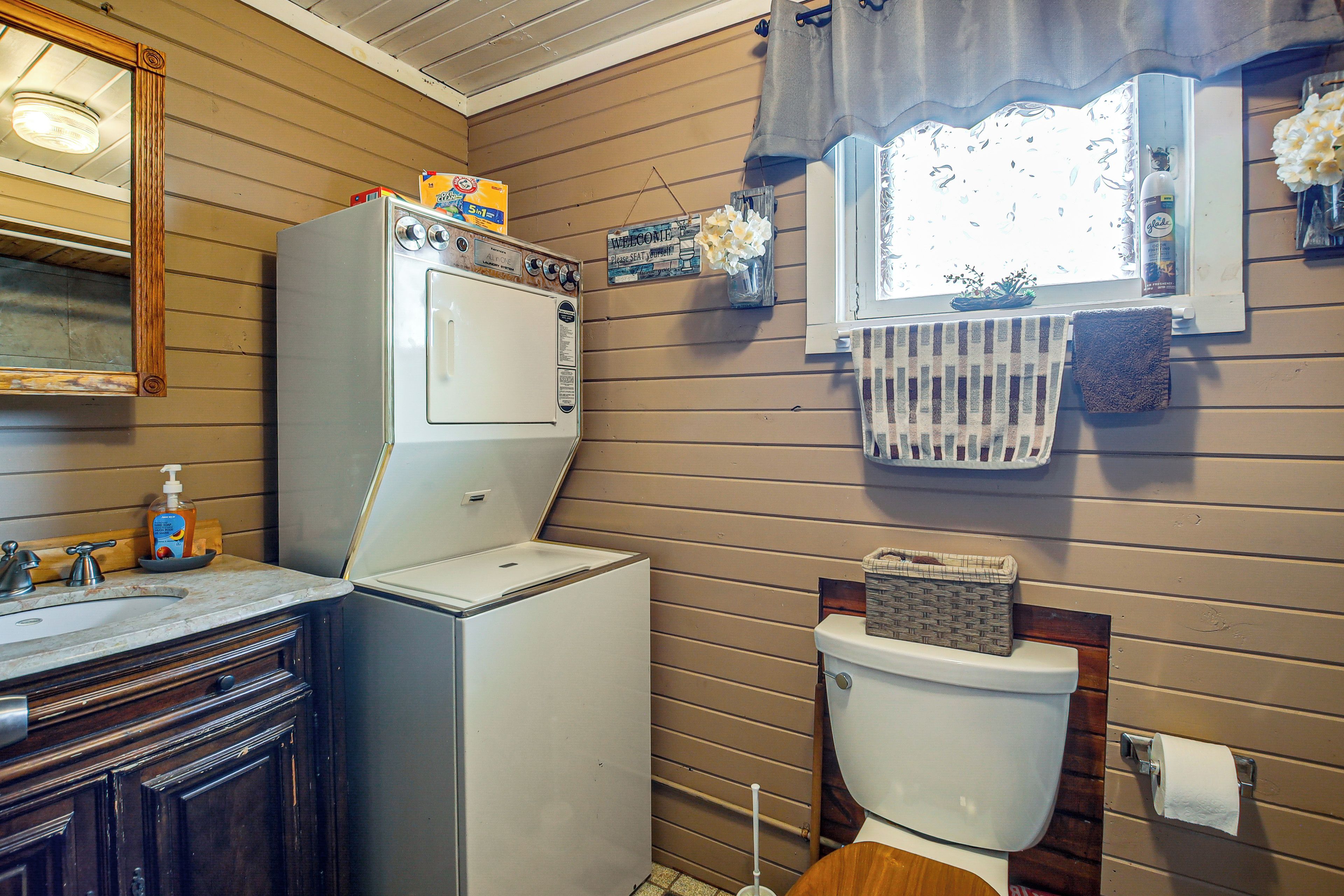 A compact bathroom with a washing machine, a toilet, a sink, and a window with curtains.