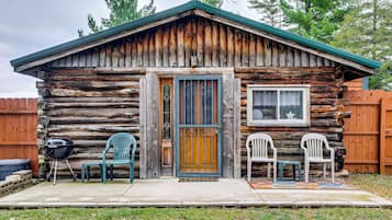 A rustic log cabin with a front door, two windows, and a porch with plastic chairs.